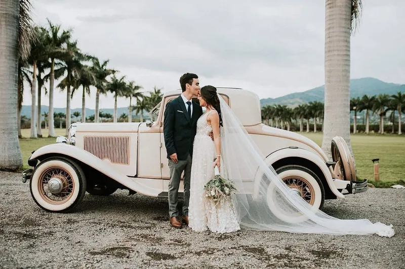 Novios junto a un auto clásico color crema, con palmeras y montañas detrás