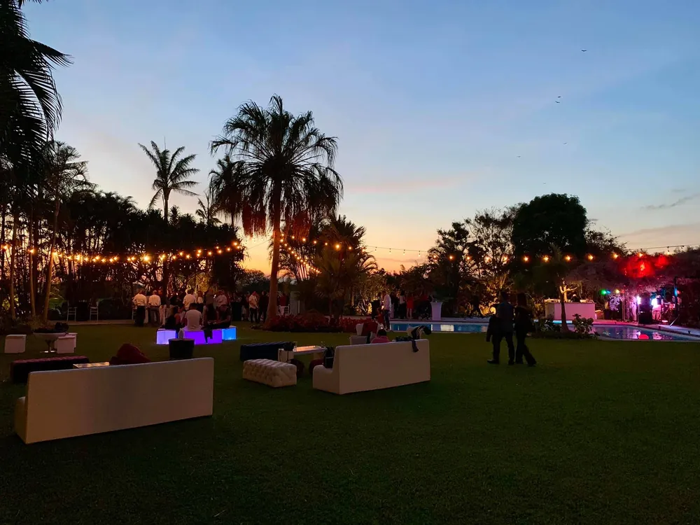 Evento al anochecer con salas blancas en el jardín y luces colgadas entre las palmeras