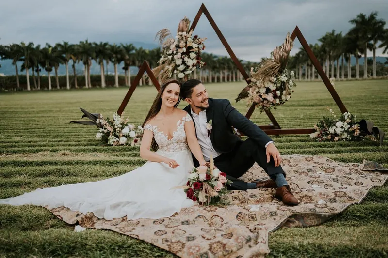 Novios sentados sobre una alfombra frente a un arco triangular de flores